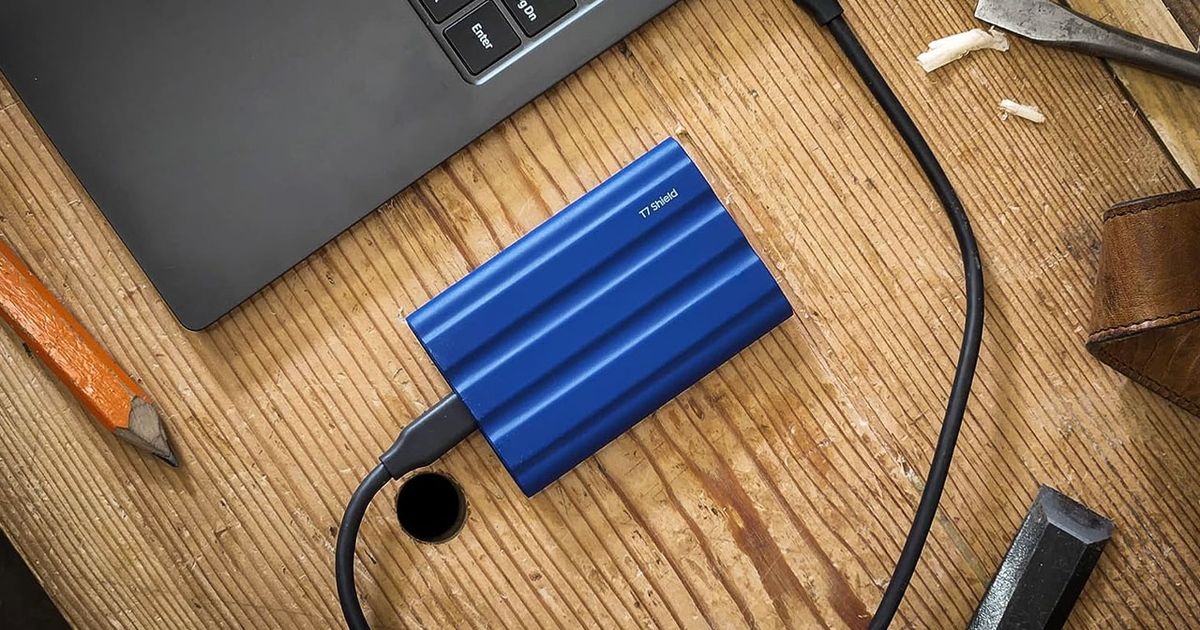 A blue hard drive connected via USB to a dark grey laptop sat on a light brown wooden desk.