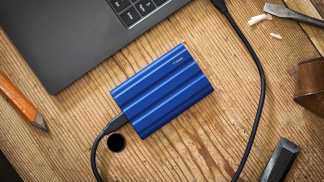 A blue hard drive connected via USB to a dark grey laptop sat on a light brown wooden desk.