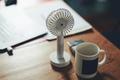 A small white fan sat on a brown desk next to paper and a white mug.