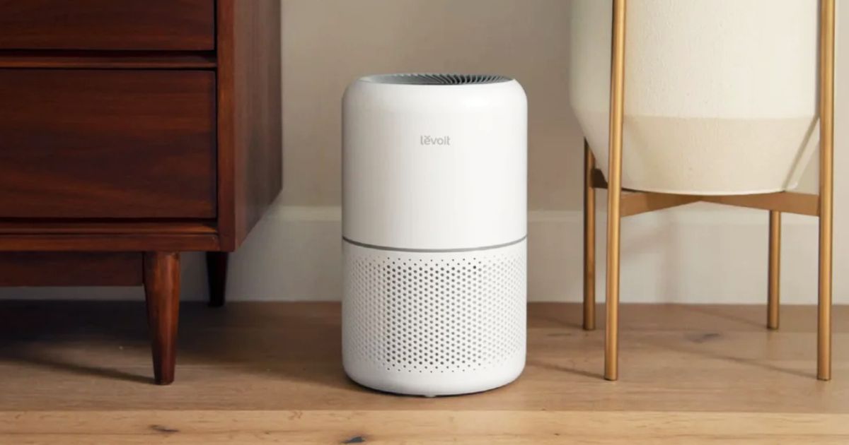 A white, cylindrical air purifier sat on a wooden floor next to a chair and a brown cabinet.