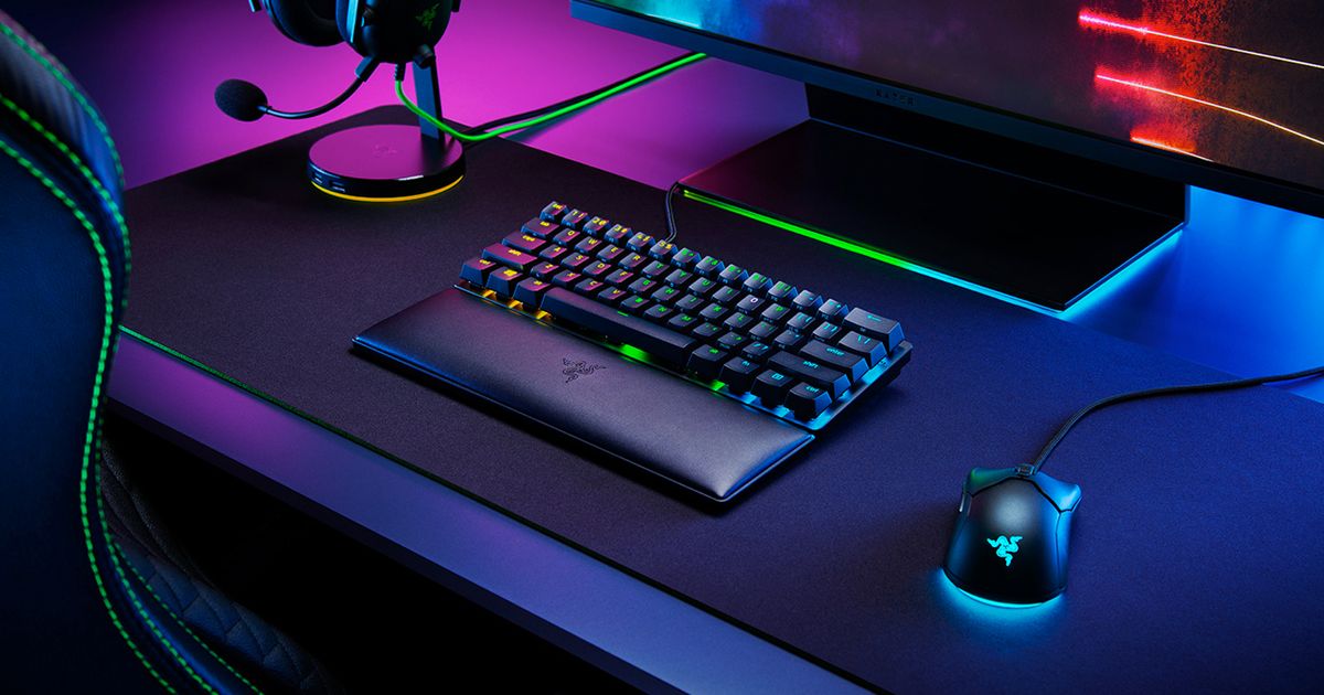 A black wrist rest in front of a black keyboard with multicoloured backlit keys, all next to a wired mouse featuring light blue lighting.