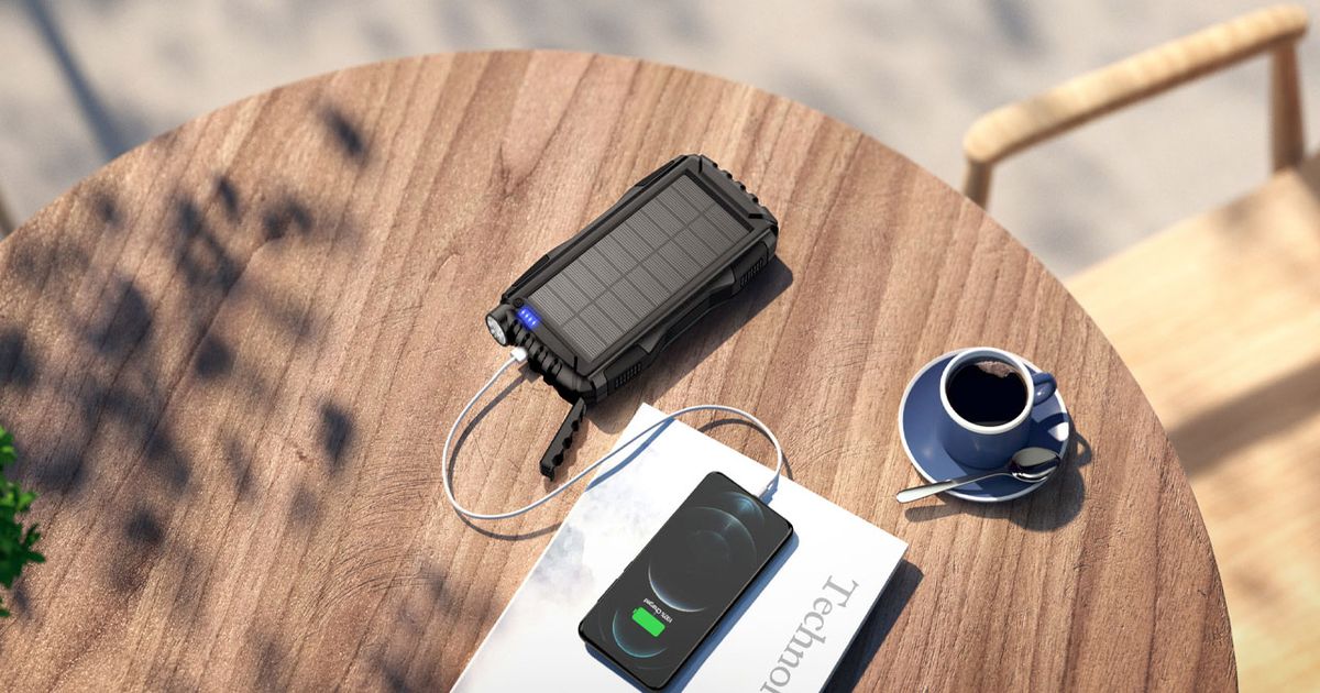 A rugged, black power bank being used to charge a black phone while sat on a brown wooden table with a blue teacup.