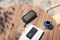 A rugged, black power bank being used to charge a black phone while sat on a brown wooden table with a blue teacup.