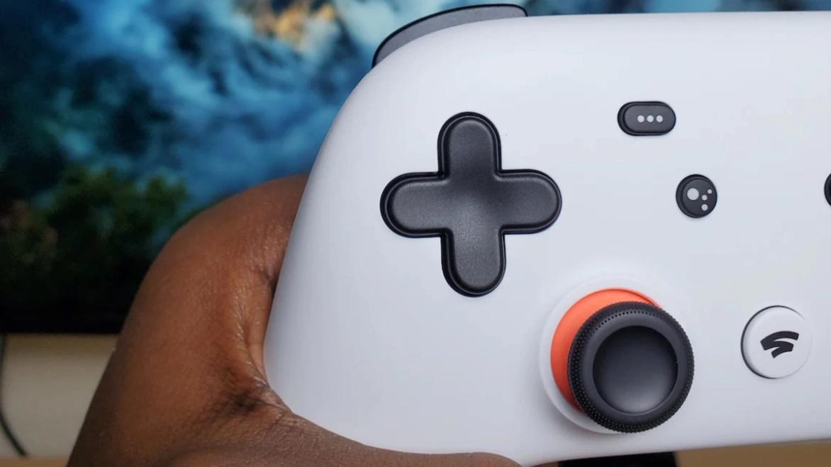 A male hand holding a Google Stadia controller in front of an out of focus TV 