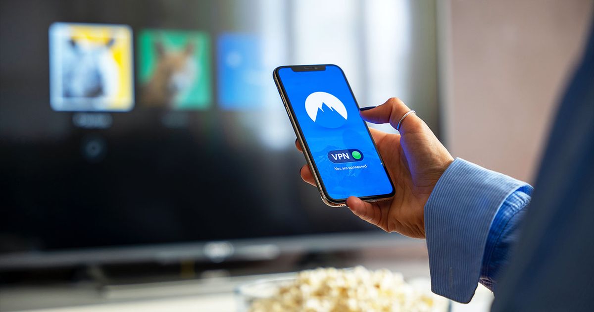 Person in a blue shirt holding a phone with a blue backdrop and a VPN on the display in front of a TV with Netflix on it.