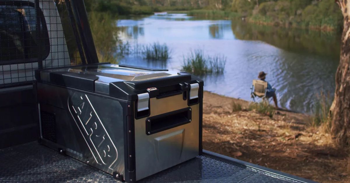 A silver and black portable fridge sat on the back of a vehicle in front of a lake.