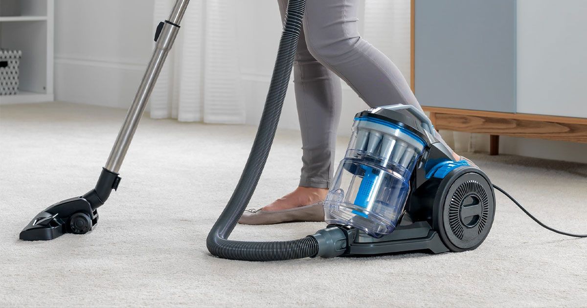 Someone in grey trousers using a small clear vacuum with blue internal components to clean a white carpet.