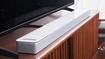 A long, thing white soundbar placed on a brown wood cabinet in front of a black TV.