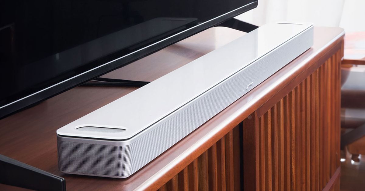 A long, thing white soundbar placed on a brown wood cabinet in front of a black TV.