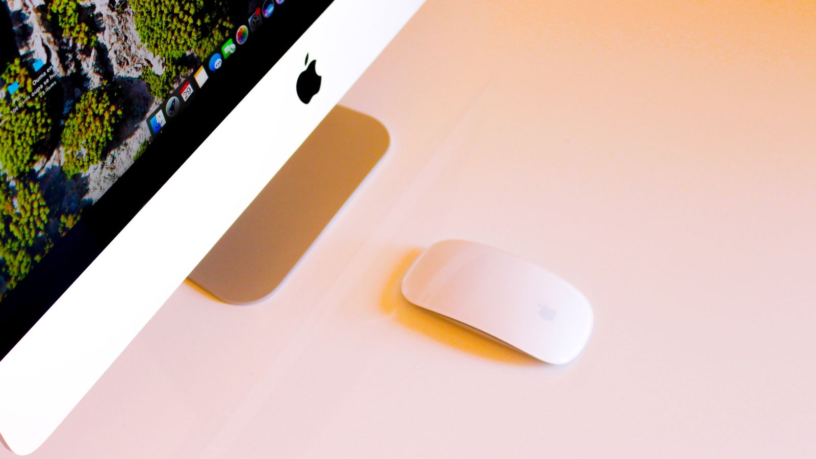 Switched on silver Mac next to a white Apple Magic Mouse placed on a light pink and orange surface.