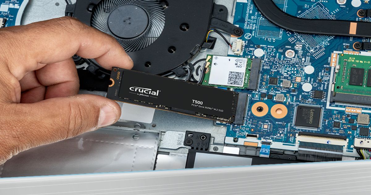 Someone holding a small, black, rectangular SSD above the inside of a PC featuring gold ends and grey branding on top.