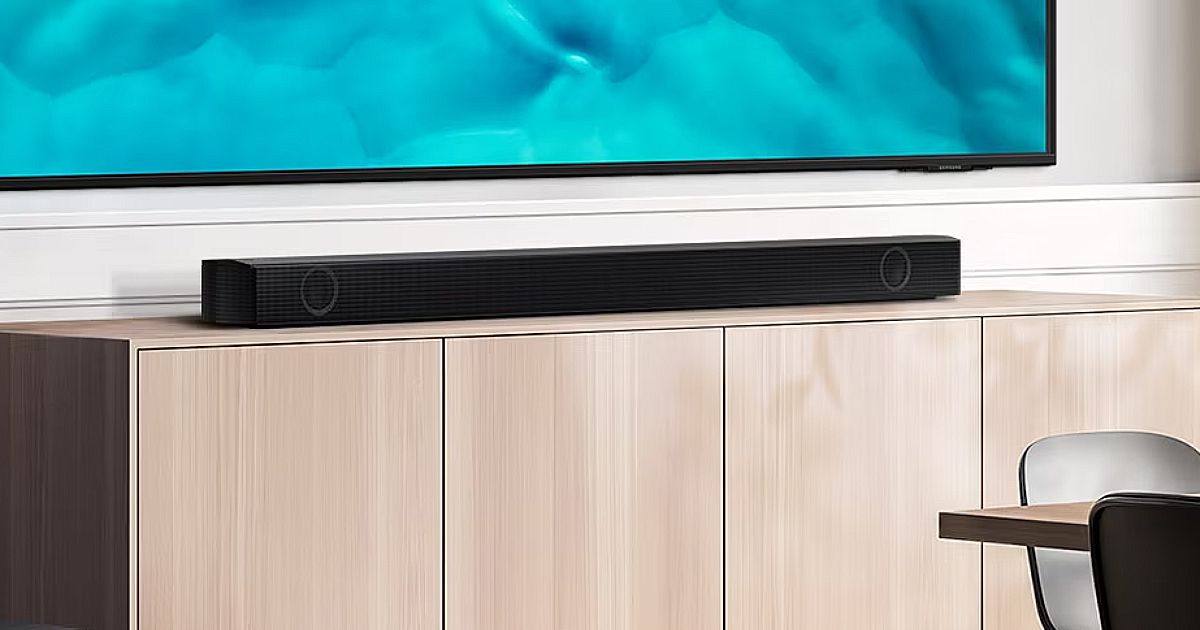 A long, black, rectangular soundbar below a large wall-mounted TV while sat on a light wood cabinet.