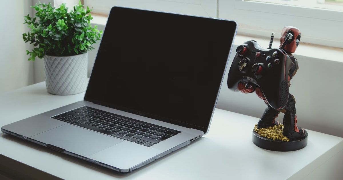A silver laptop open on a desk next to a figure of Deadpool holding an Xbox controller.