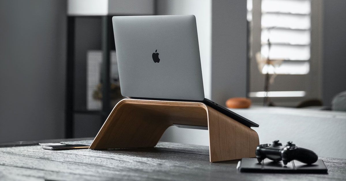 A dark grey MacBook sat on top of a brown laptop stand with a phone to one side and a black controller the other.