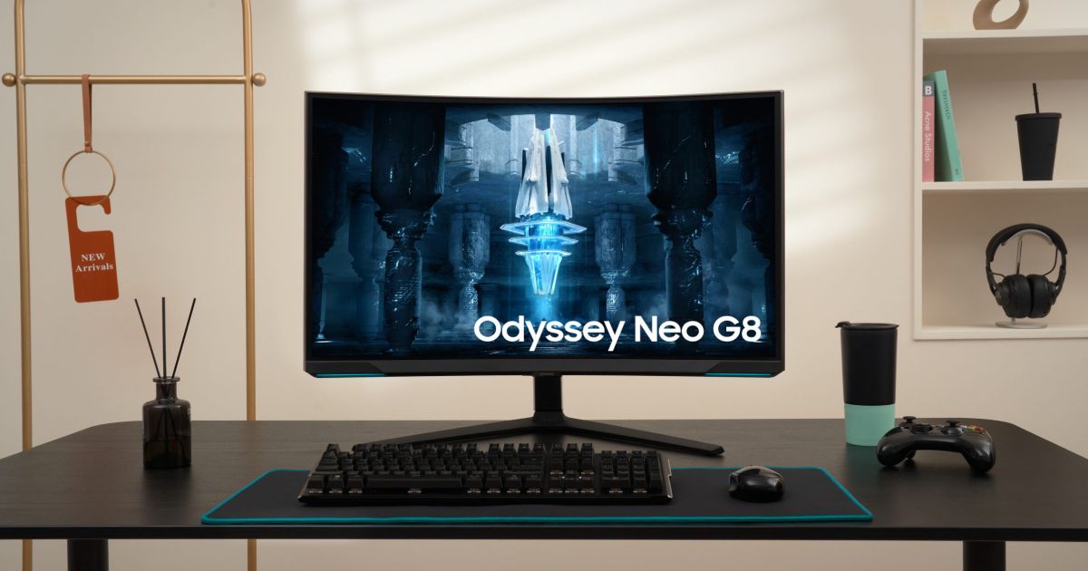 A black Samsung monitor with Odyssey Neo G8 branding on the display sat on a desk with a keyboard, mouse, and controller.