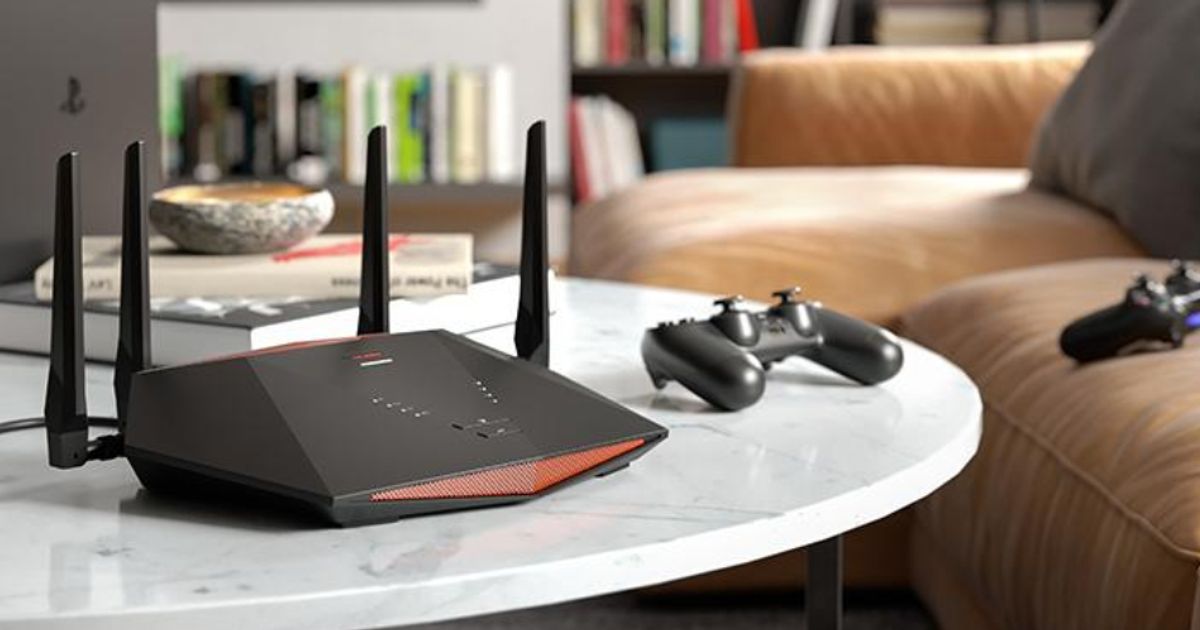 A black WiFi router with red trim at the front and back sat on a white table next to a black PlayStation controller.
