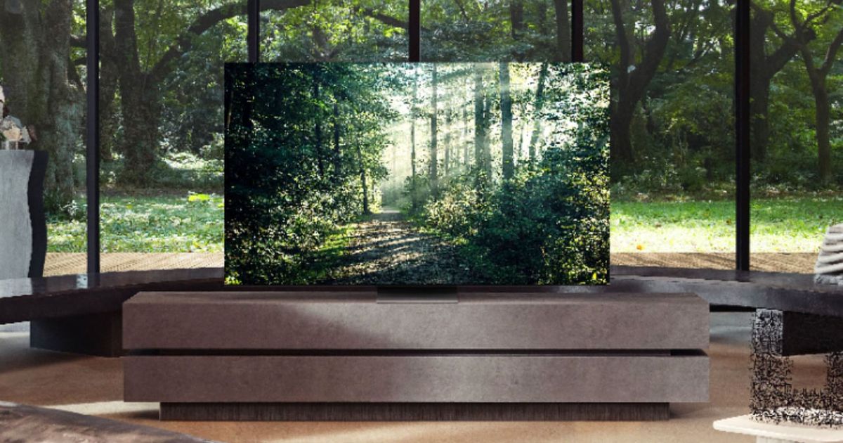 A flatscreen TV with a forest scene on the display sat on a brown stand in front of a window looking out onto a forest.