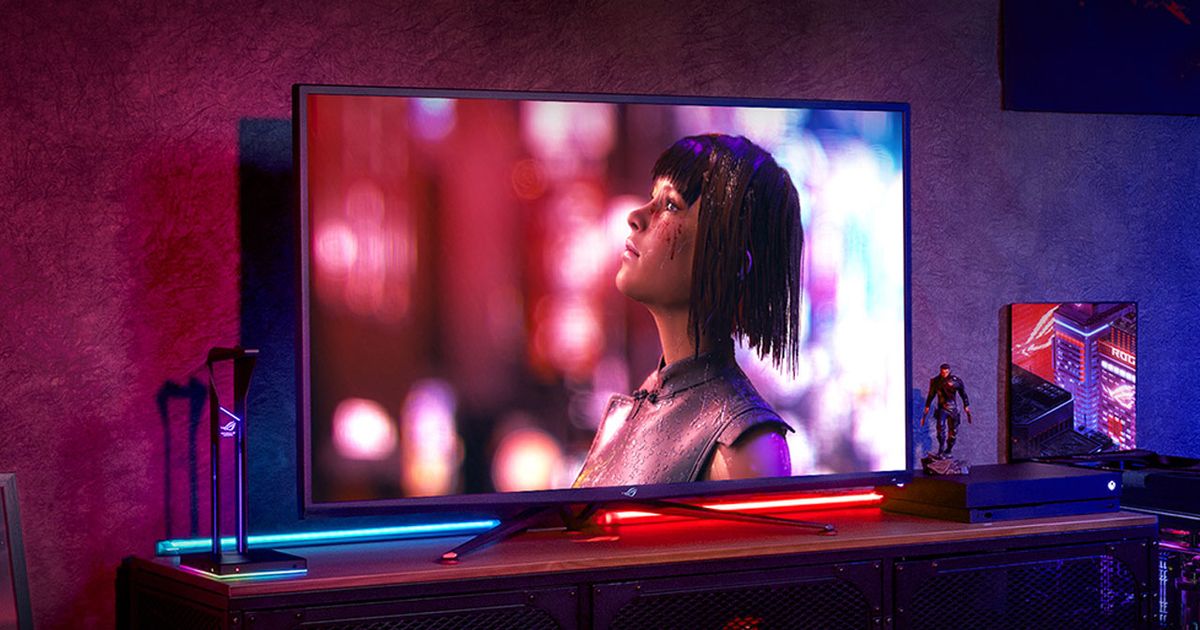 A large gaming monitor with a video game character looking up on the display sat in a red and blue-lit room next to a PlayStation.