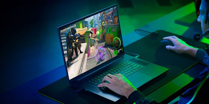 Image of someone in a darkerned room featuring blue and green lighting playing Overwatch on a gaming laptop.