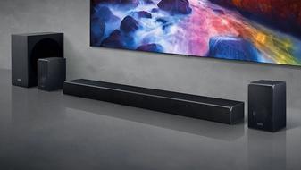 A long black soundbar next to black speakers and a subwoofer in a grey room. The speaker setup is below a wall-mounted TV.