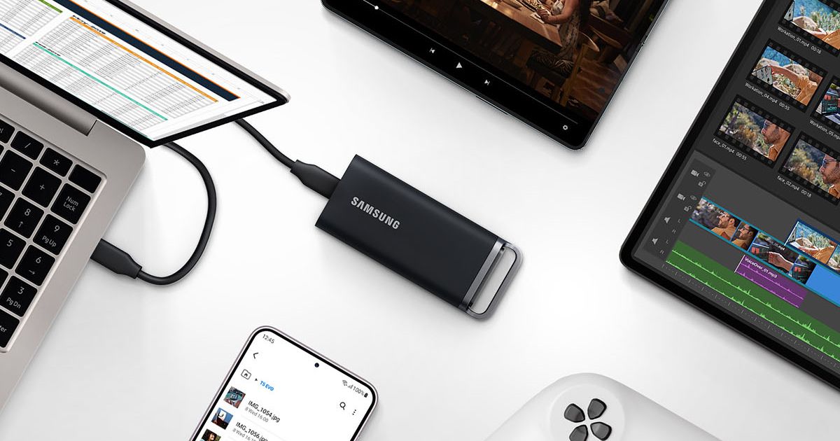 A black portable SSD connected by a wire to a silver laptop while surrounded by other tech.