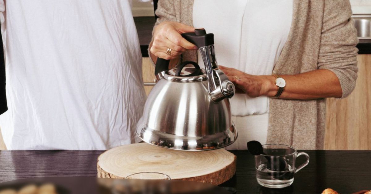 Someone in white top and beige cardigan lifting a silver kettle off a wooden plate, pouring into a glass mug.