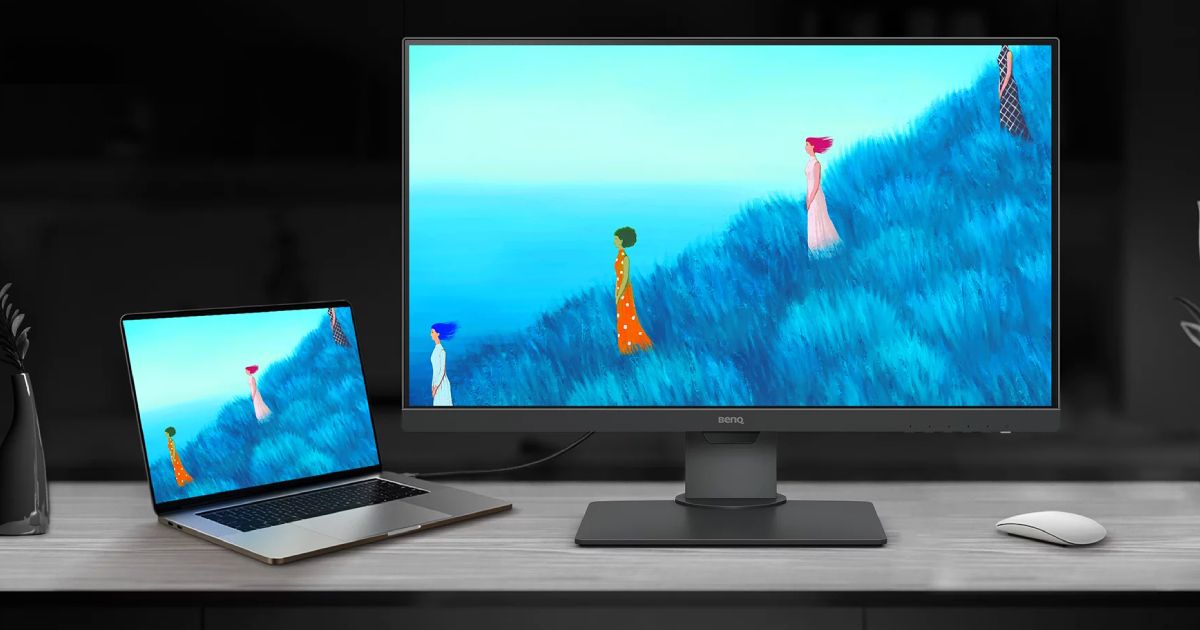 A black monitor with artwork of four women standing on a blue grass hillside on the display next to a MacBook.
