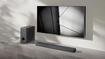 A dark grey soundbar placed in a grey and white room next to a speaker and below a flatscreen TV mounted to a wall.