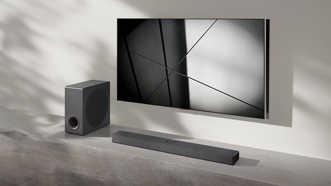 A dark grey soundbar placed in a grey and white room next to a speaker and below a flatscreen TV mounted to a wall.
