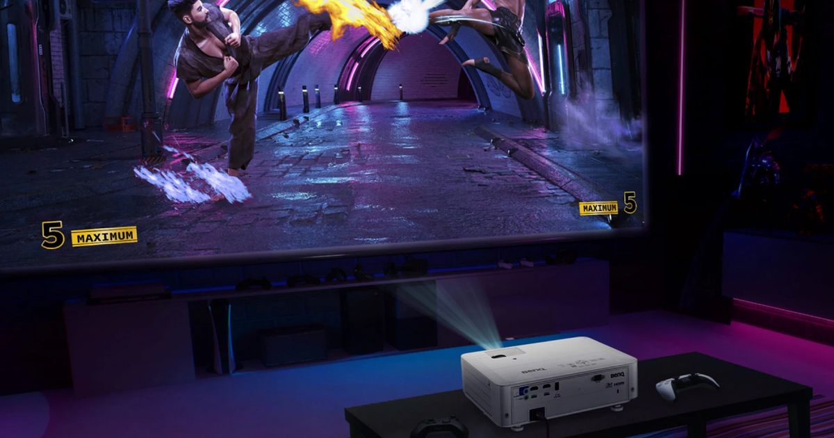 A white projector on a table next to a PS5 controller projecting a fighting game onto a large screen.