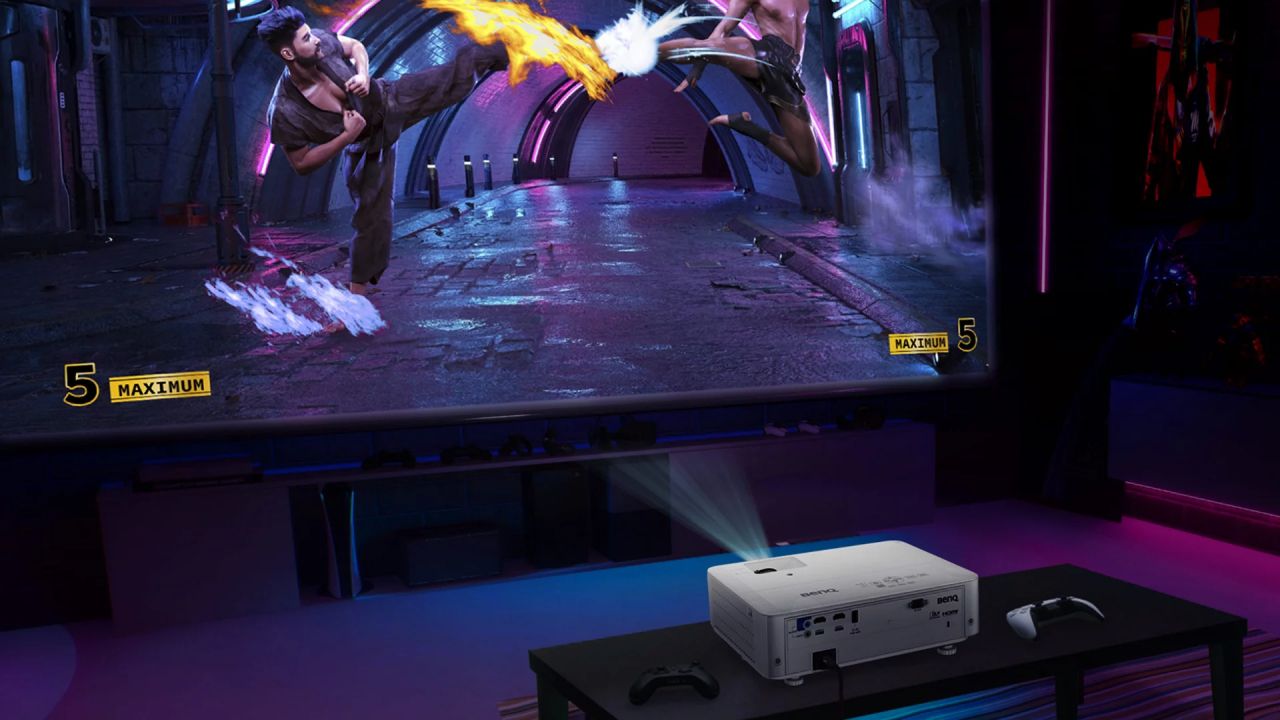 A white projector on a table next to a PS5 controller projecting a fighting game onto a large screen.