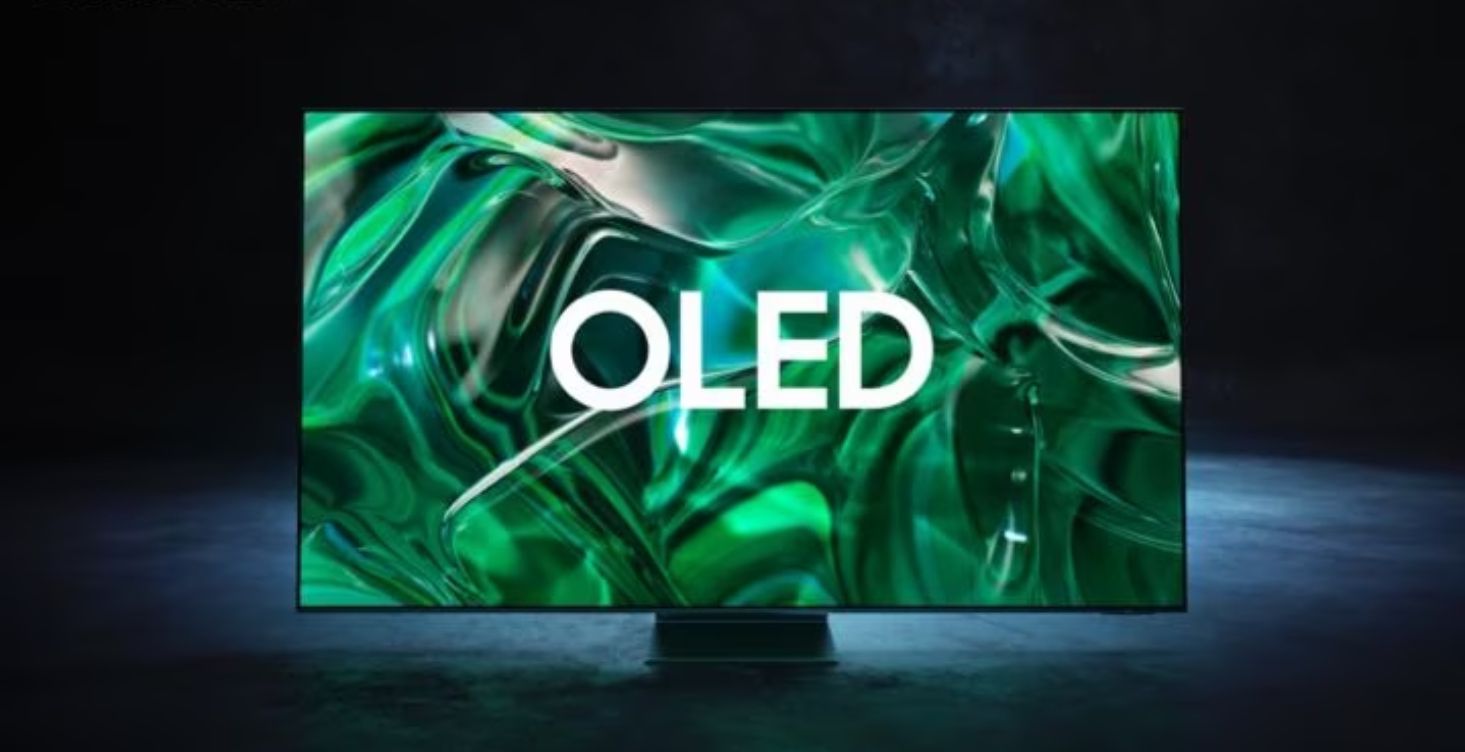 Image of a large flatscreen Samsung TV featuring OLED in white text on the display in front of a green backdrop.